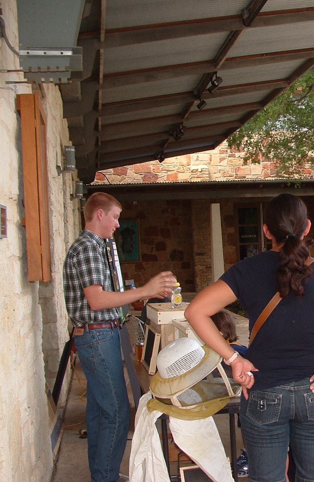 A teenage beekeeper in a plaid shirt holds up a jar of honey beside an observation hive, talking to visitors under a stone building's overhang.