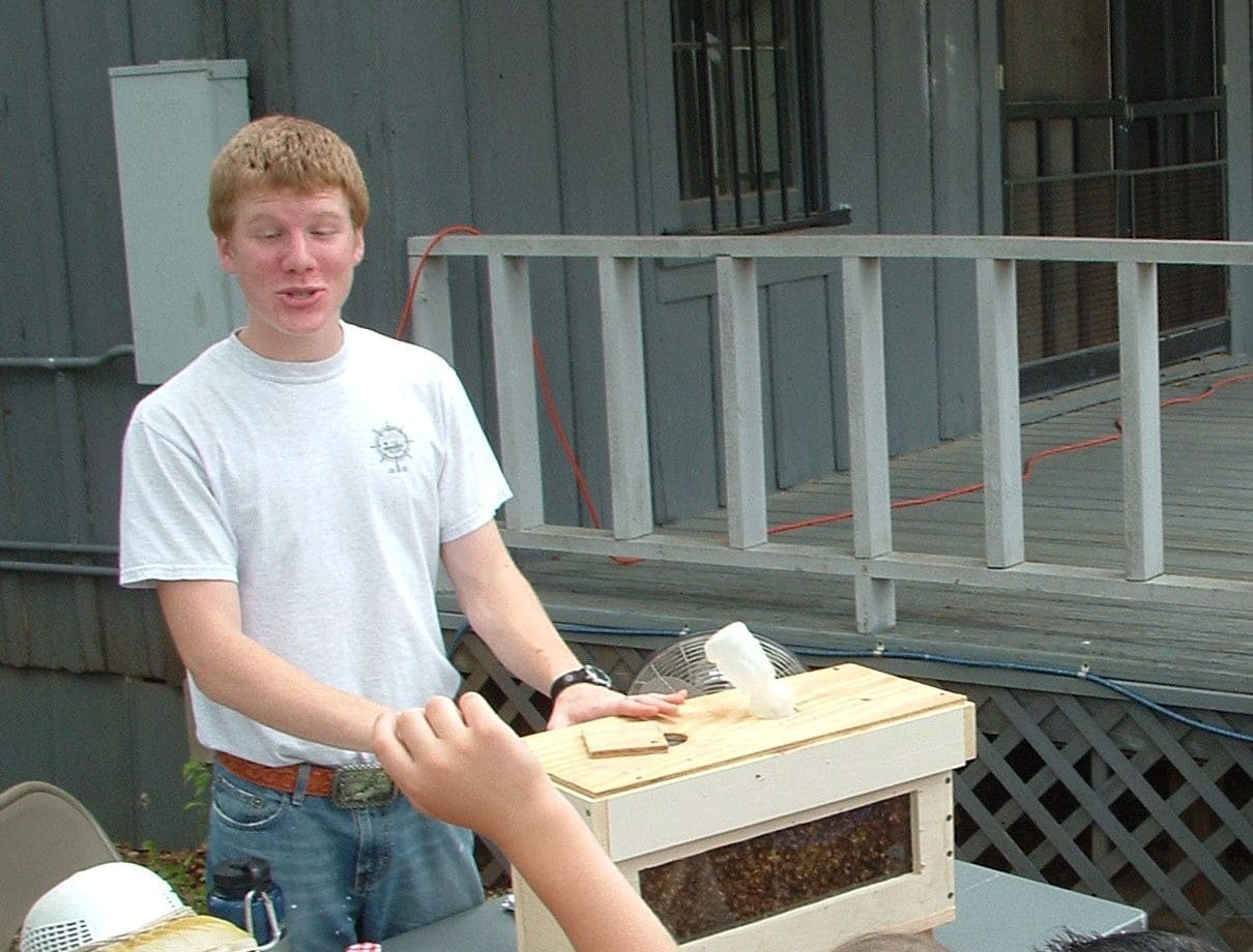 A teenage beekeeper stands behind a wooden observation hive with glass sides, talking to visitors at an outdoor event.