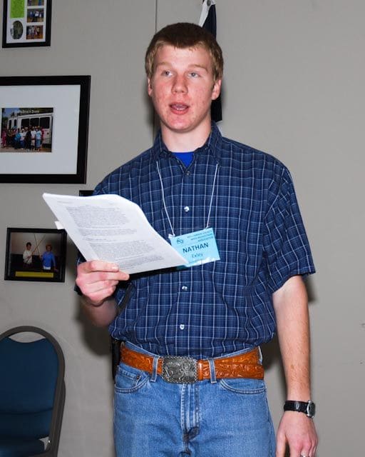 A teenage Nathan Exley, wearing a name badge, reads aloud from a sheet of paper at a beekeepers' meeting.